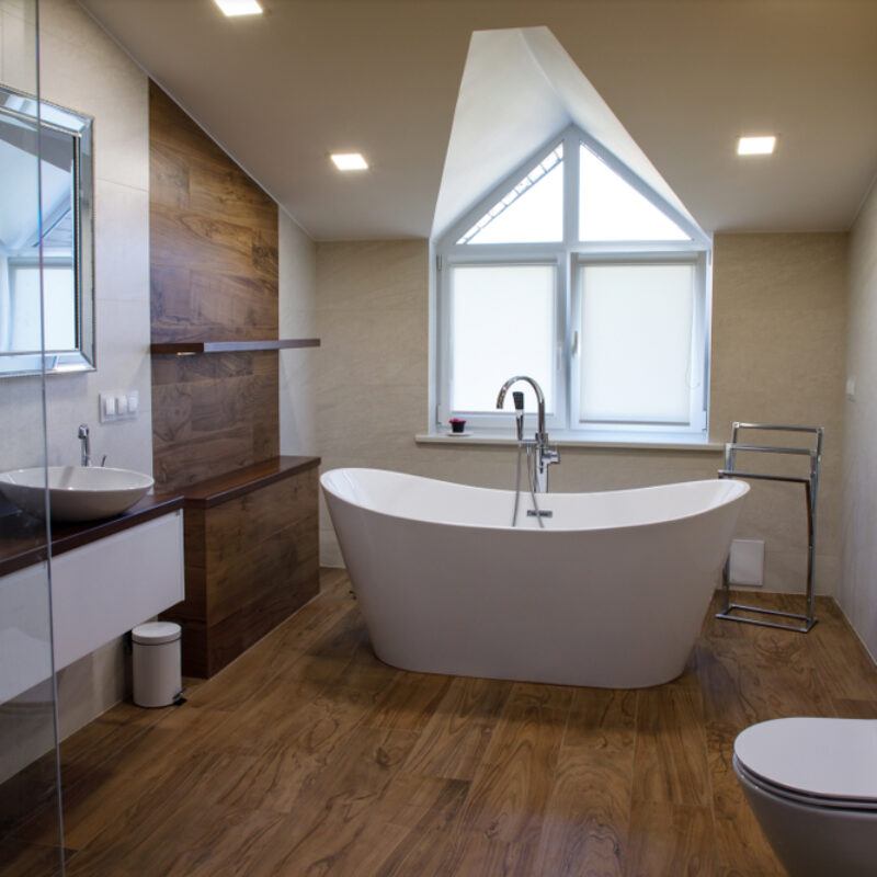 Bathroom installations in Northamptonshire. A modern bathroom with free standing bath in front of a window. The bathroom has waterproof wooden flooring, two sinks and is bright and airy. The ceiling slopes down to the window and has square spotlights embedded in.
